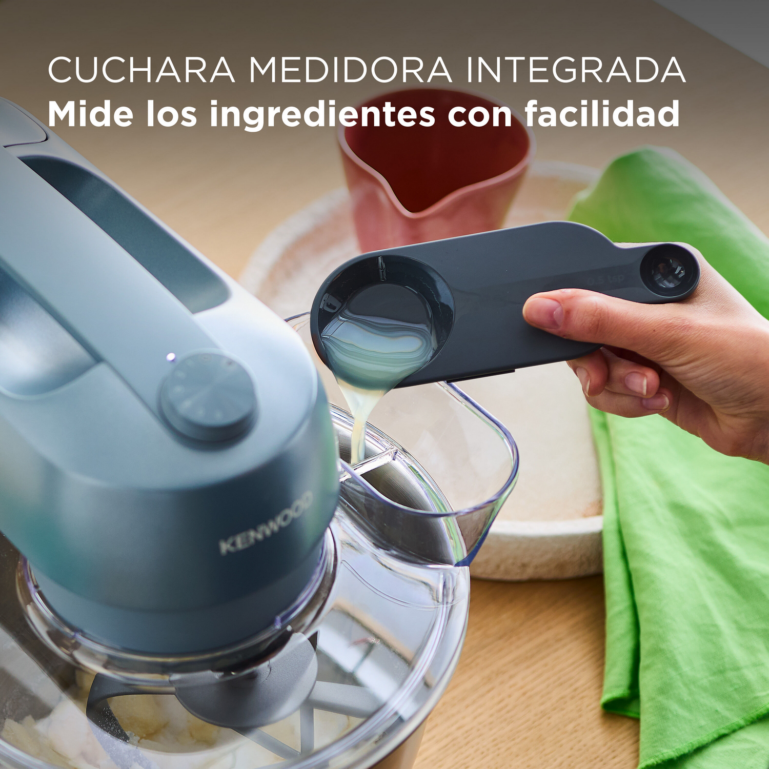 Una persona usa una batidora Kenwood azul, vertiendo líquido de la cuchara medidora en la batidora.