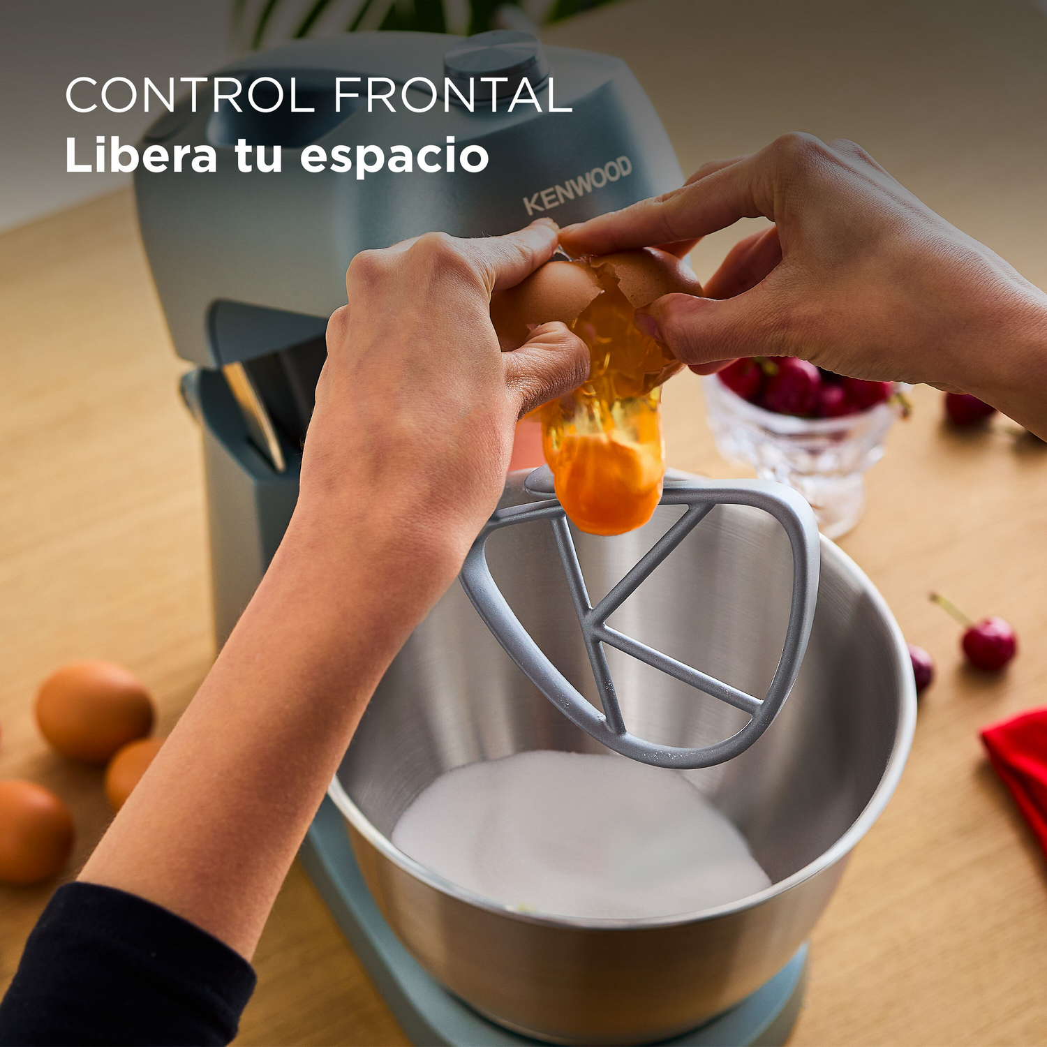 Una persona rompiendo un huevo en un bol con azúcar. Una batidora Kenwood está en el fondo. Hay cerezas en la mesa.