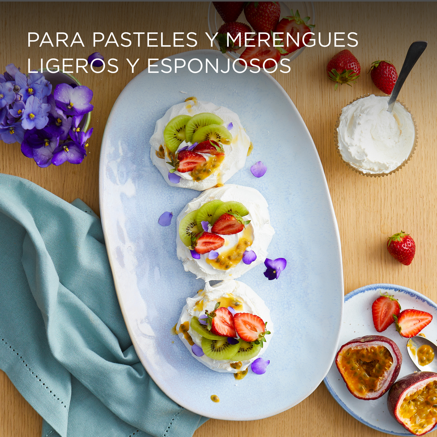 Pavlovas con kiwi, fresas y maracuyá. Están en un plato azul, con violetas y un paño azul.