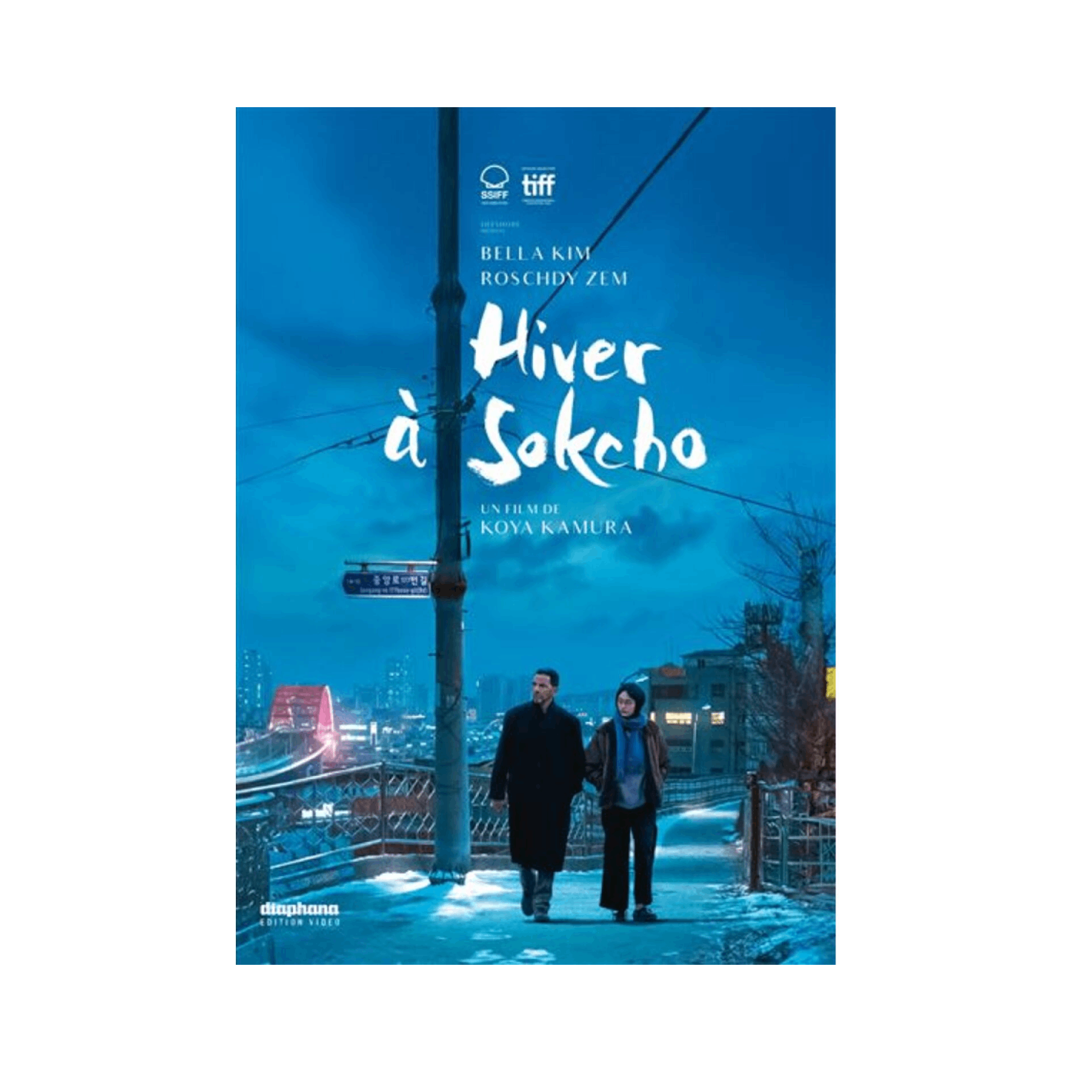 Affiche pour 'Hiver à Sokcho'. Deux personnes marchant dans une rue enneigée, lumières de la ville et bâtiments.