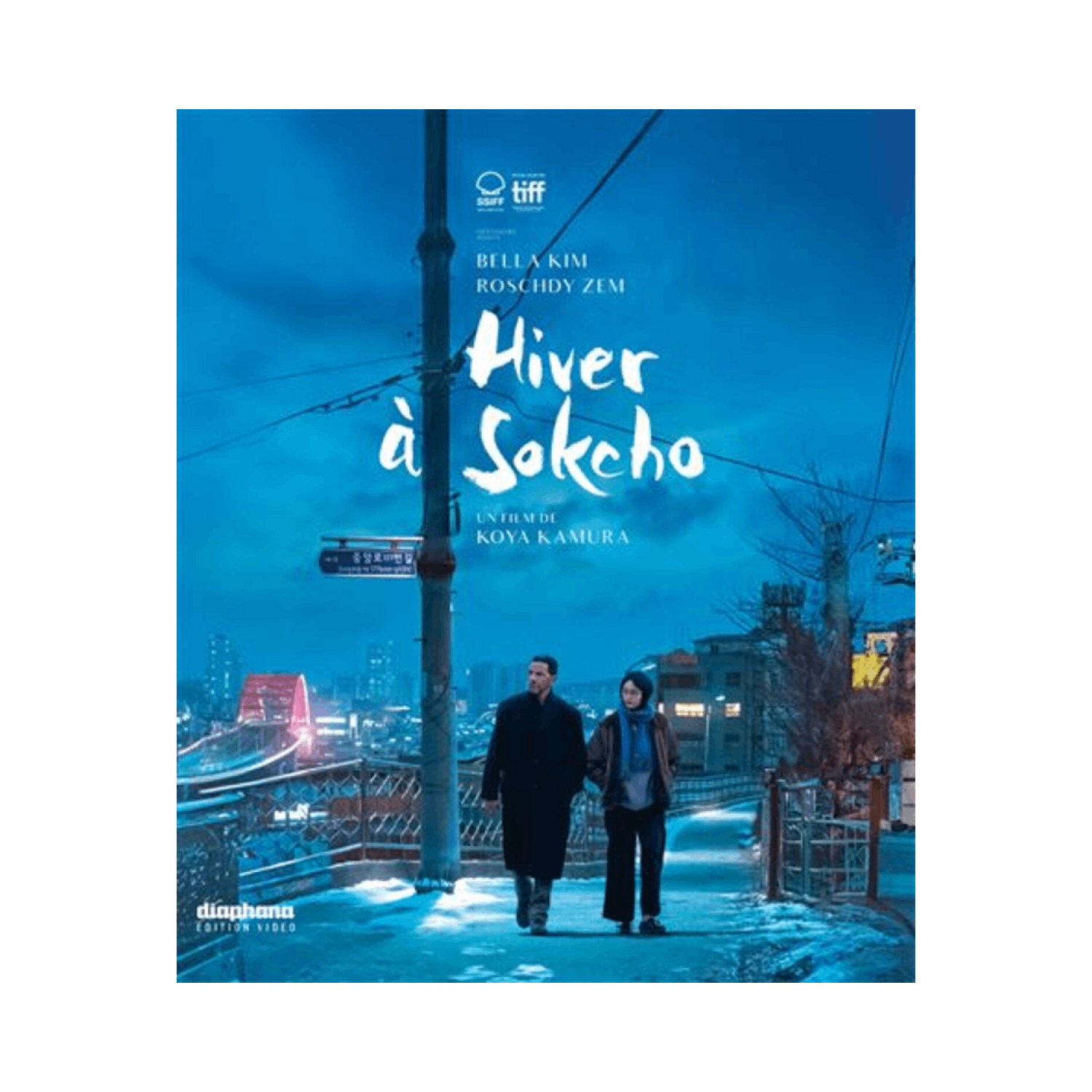 Affiche pour 'Hiver à Sokcho'. Deux personnes marchant dans une rue enneigée, lumières de la ville et bâtiments.