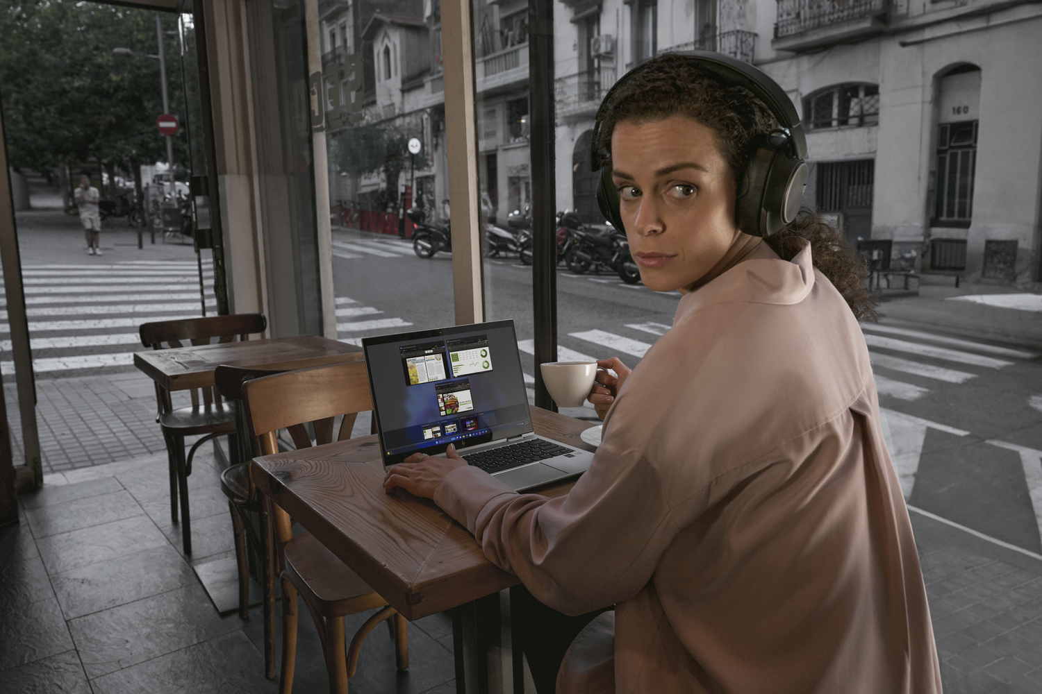 Una mujer con auriculares usa una laptop en una mesa de café, mirando a la cámara.
