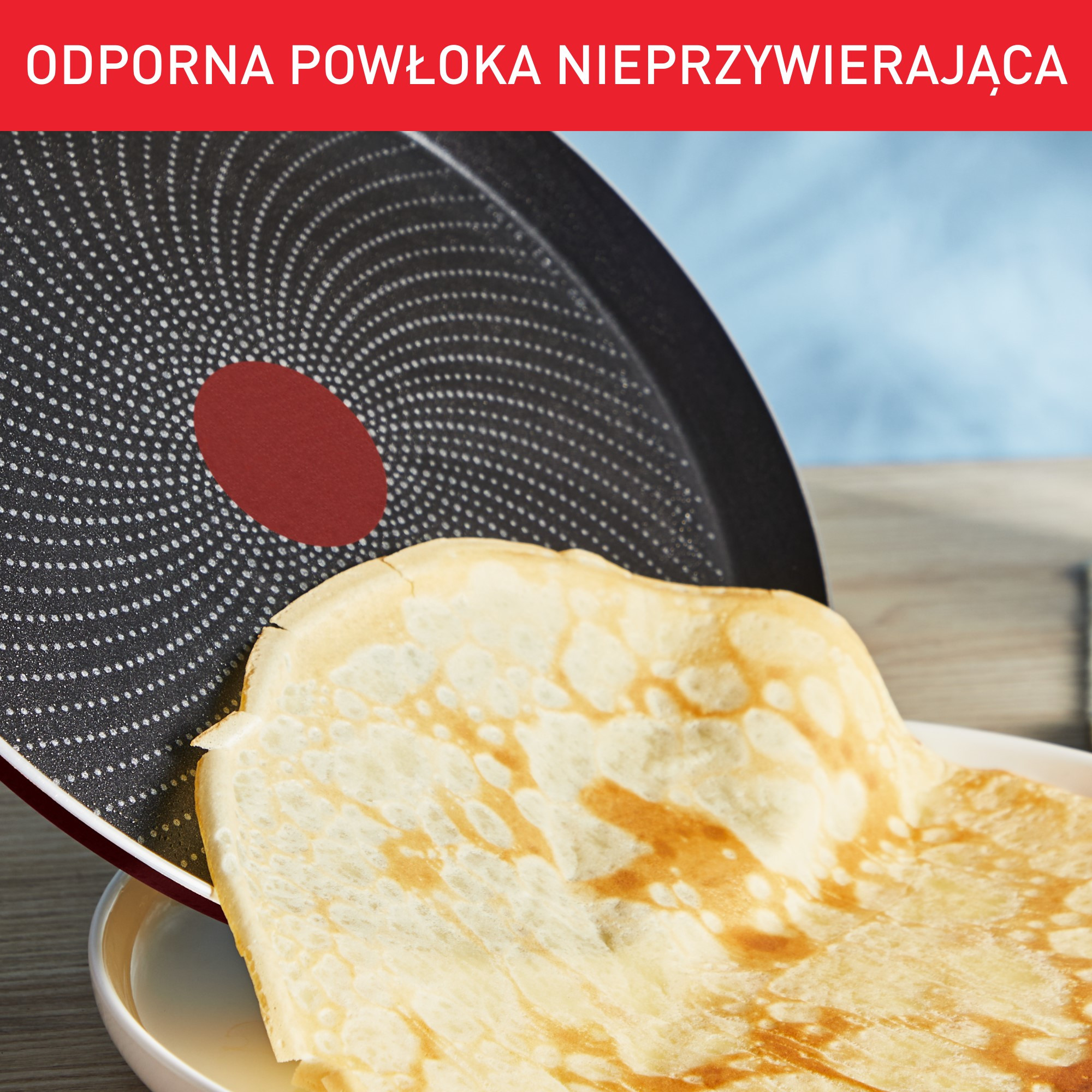 Naleśnik obracany z patelni nieprzywierającej. Patelnia jest czarna i czerwona. Naleśnik jest złoty.