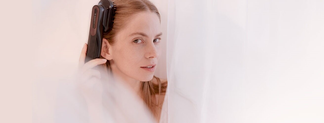 Kobieta z długimi włosami używa urządzenia do wzrostu włosów. Jest przed białą zasłoną, patrzy w kamerę.