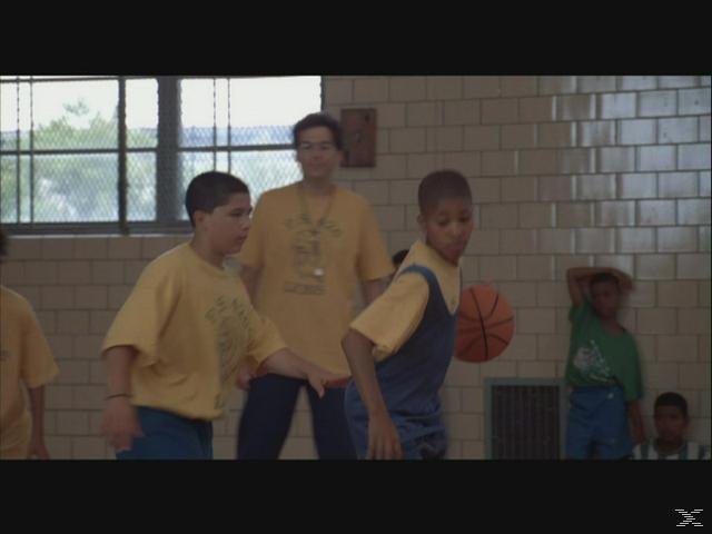 Jungen spielen Basketball in einer Turnhalle. Ein Trainer ist im Hintergrund.