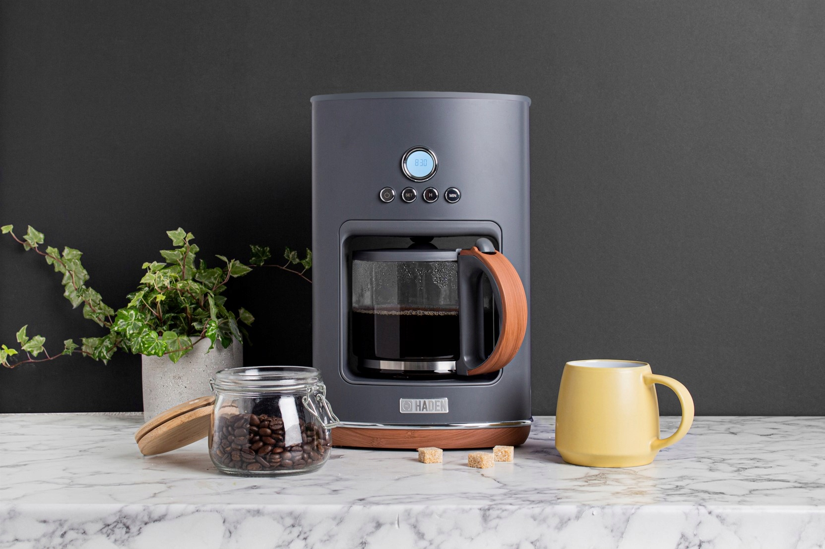Ciemnoszary ekspres do kawy z ziarnami kawy, żółtym kubkiem i kostkami cukru na marmurowym blacie.
