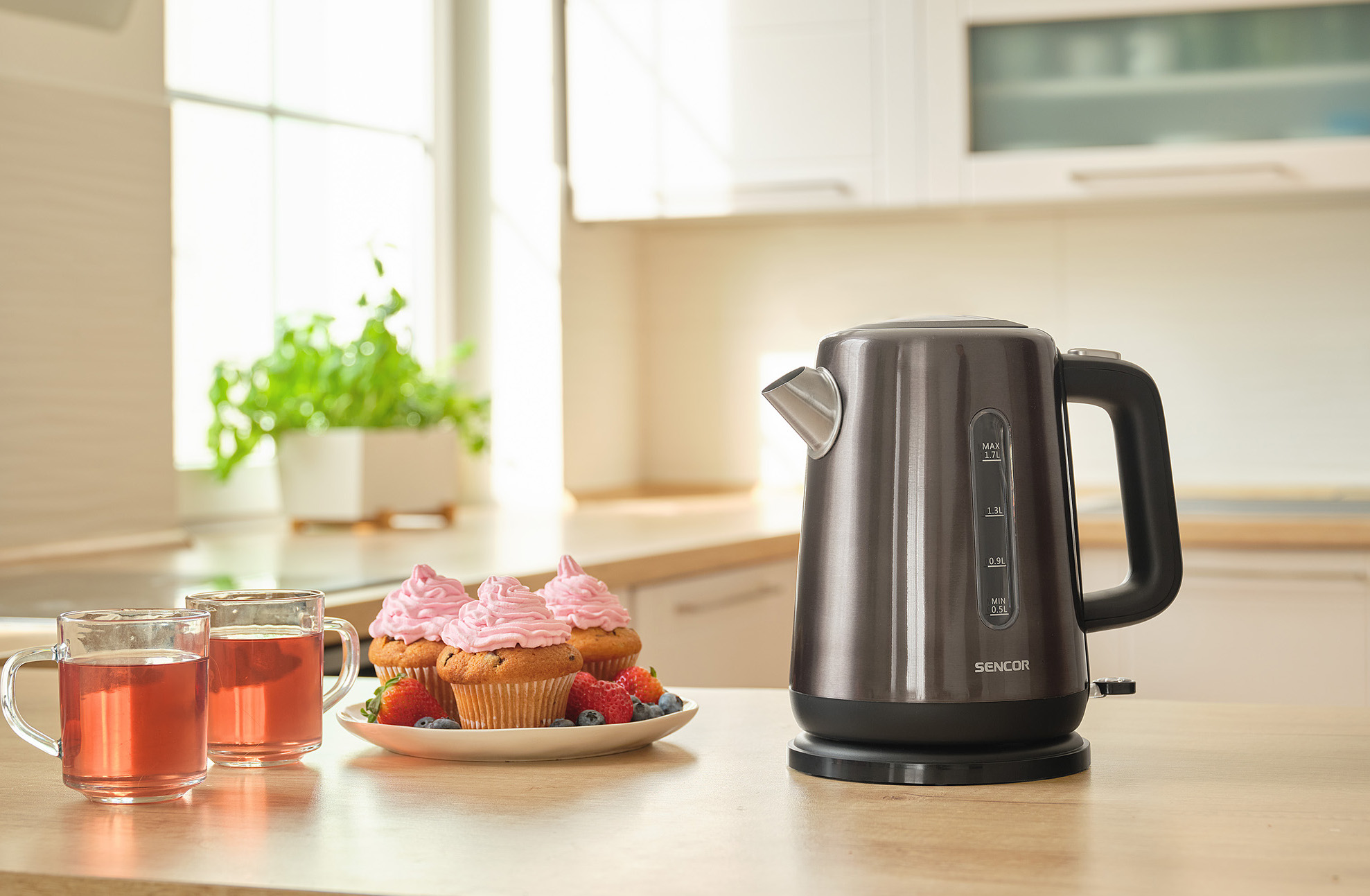 Czarny czajnik na drewnianym stole z babeczkami, napojami i widocznym tłem kuchni.