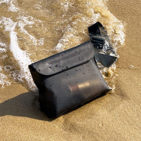 Czarna wodoodporna torba na mokrym piasku, częściowo zanurzona w wodzie. Otaczają ją fale. Czarny pasek przymocowany do torby.