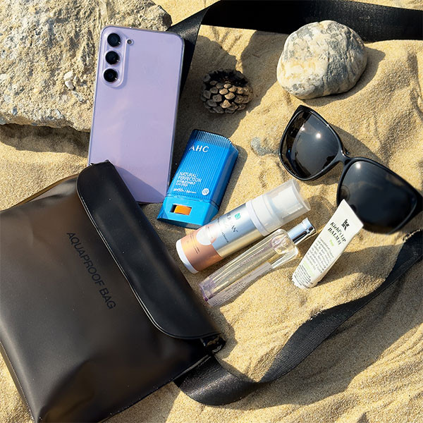 Scena na plaży: telefon, krem ​​z filtrem, okulary przeciwsłoneczne, torba i kosmetyki na piasku. Tło ma skałę, szyszkę. Wszystko w słoneczny dzień.