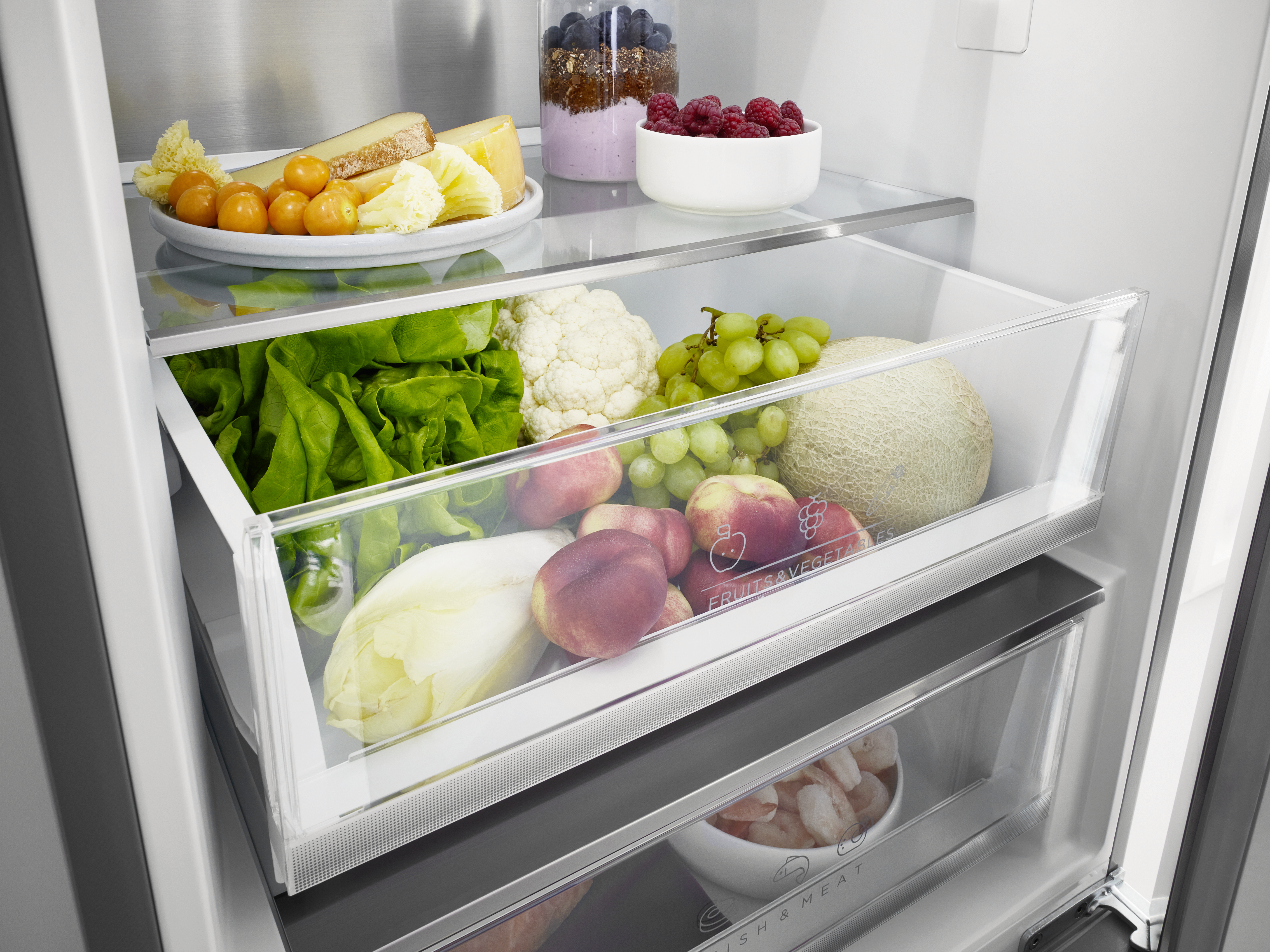 Kühlschrankinnenraum mit Obst und Gemüse in Schubladen. Oberes Regal hat Käse, Beeren. Glas mit Beeren.