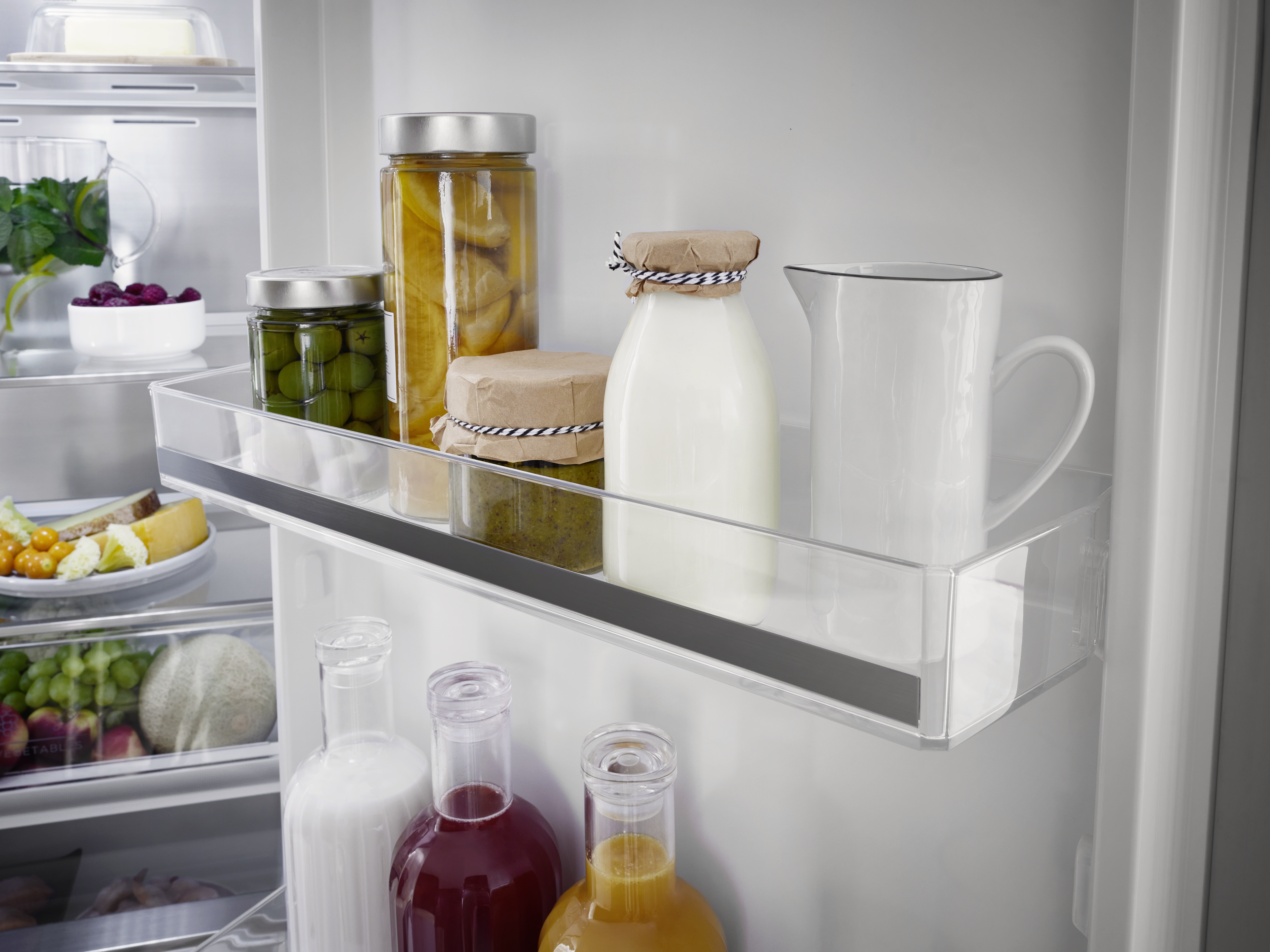 Kühlschrankinnenraum mit Gläsern, Milchflaschen und einem Krug. Gläser enthalten Oliven, Pfirsiche und Pesto. Milch- und Saftflaschen.