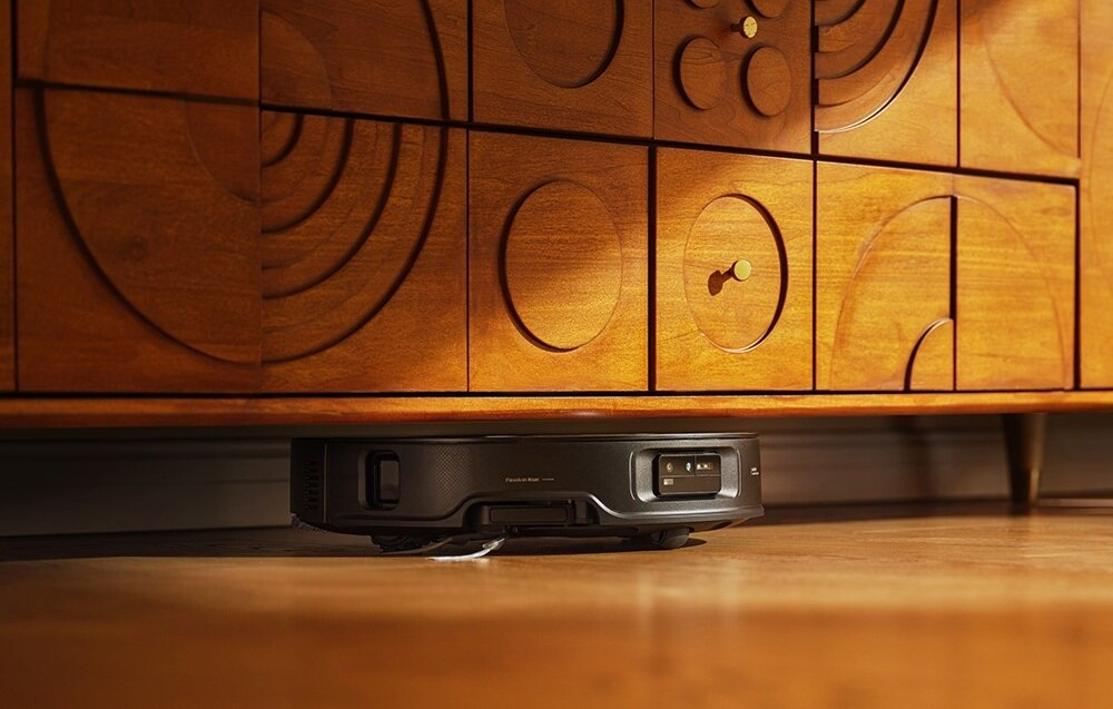 Robot odkurzający pod drewnianą szafką. Szafka ma dekoracyjny wzór, a podłoga jest drewniana.