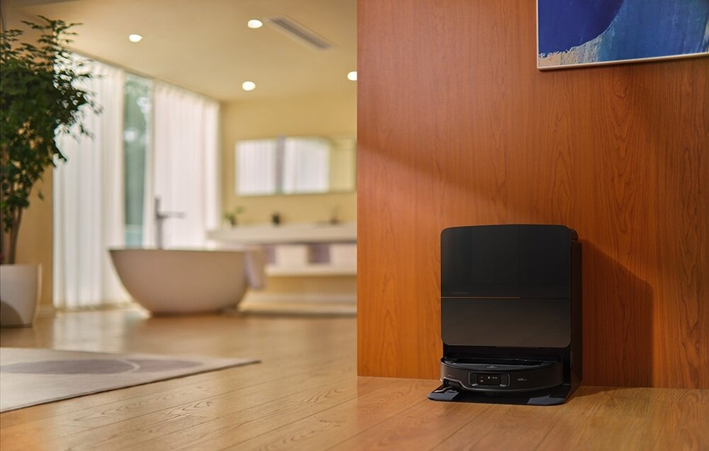 Czarny robot odkurzający stoi przy drewnianej ścianie. W tle łazienka z wanną.