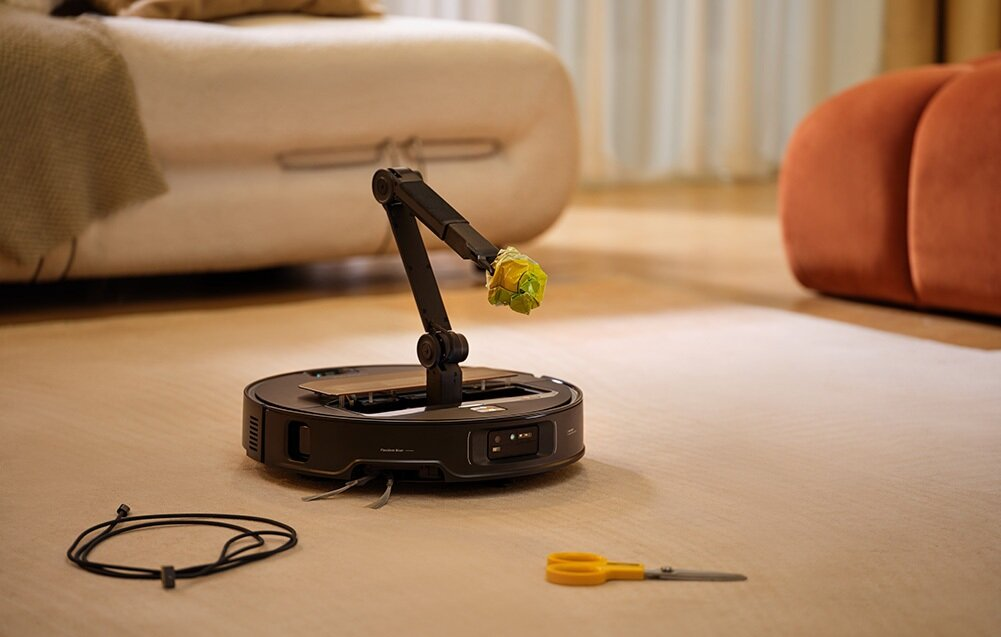 Robot odkurzający z ramieniem trzymającym zielony przedmiot. Nożyczki i czarny kabel na beżowej podłodze. Kanapa w tle.