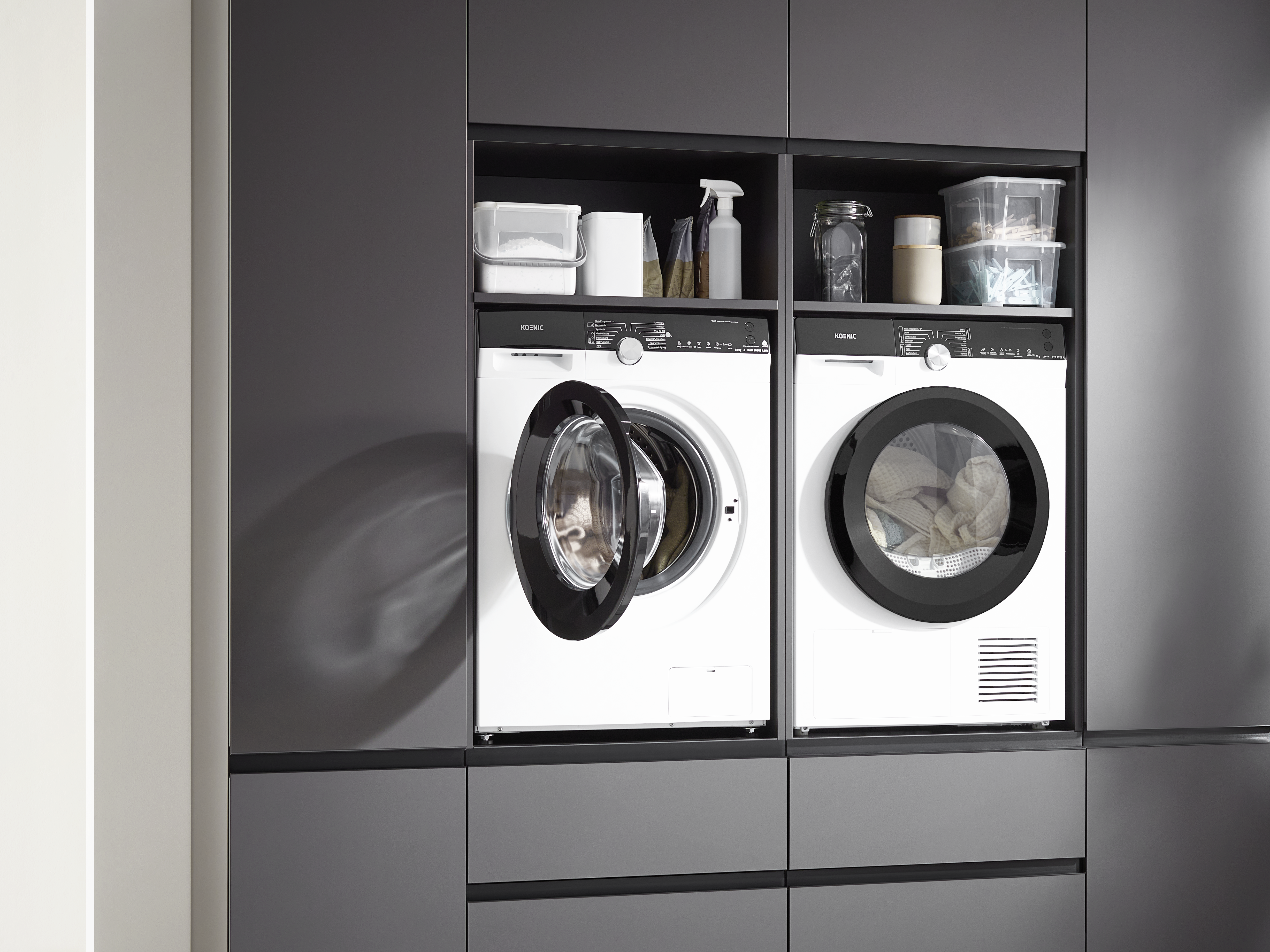 Zwei weiße Waschmaschinen mit geöffneten Türen in einem Einbauschrank. Regale darüber enthalten Reinigungsmittel und Behälter.