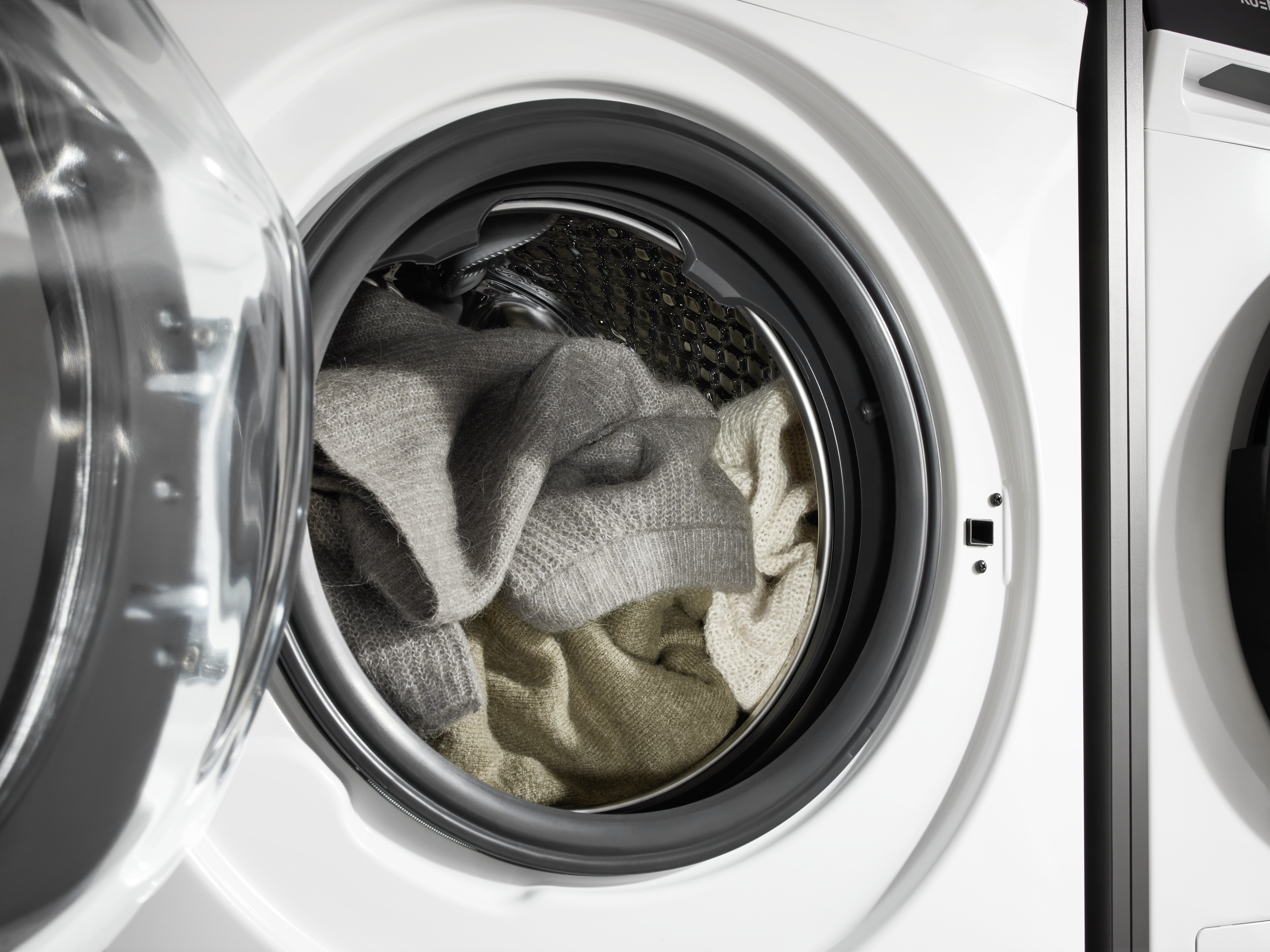 Waschmaschine mit offener Tür. Kleidung darin: graue und beige Pullover. Weiße Maschine mit schwarzem Rand. Glänzende Glastür.