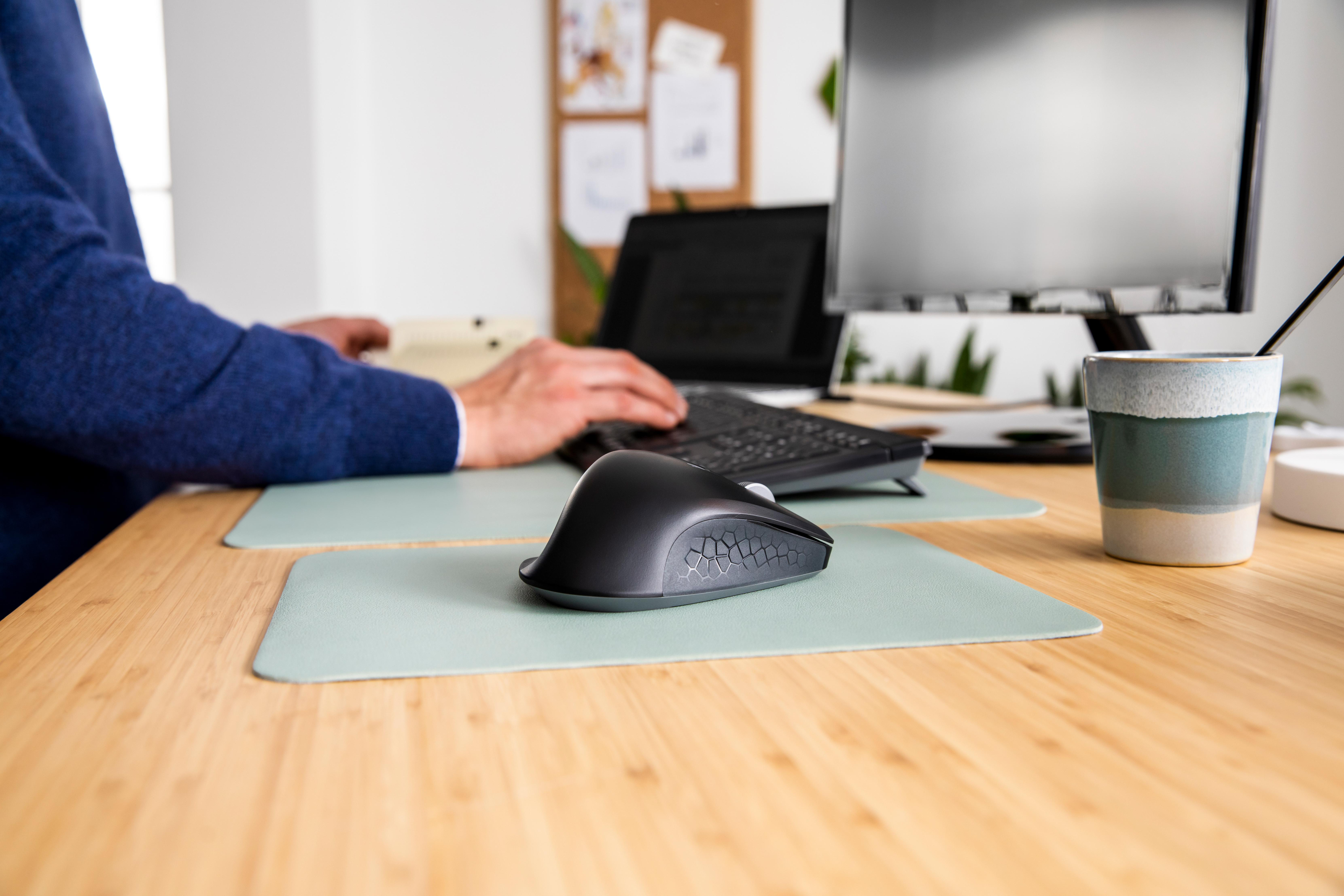 Eine Person tippt auf einer Tastatur an einem Schreibtisch. Maus, Monitor und Becher sind sichtbar. Der Schreibtisch ist aus Holz.