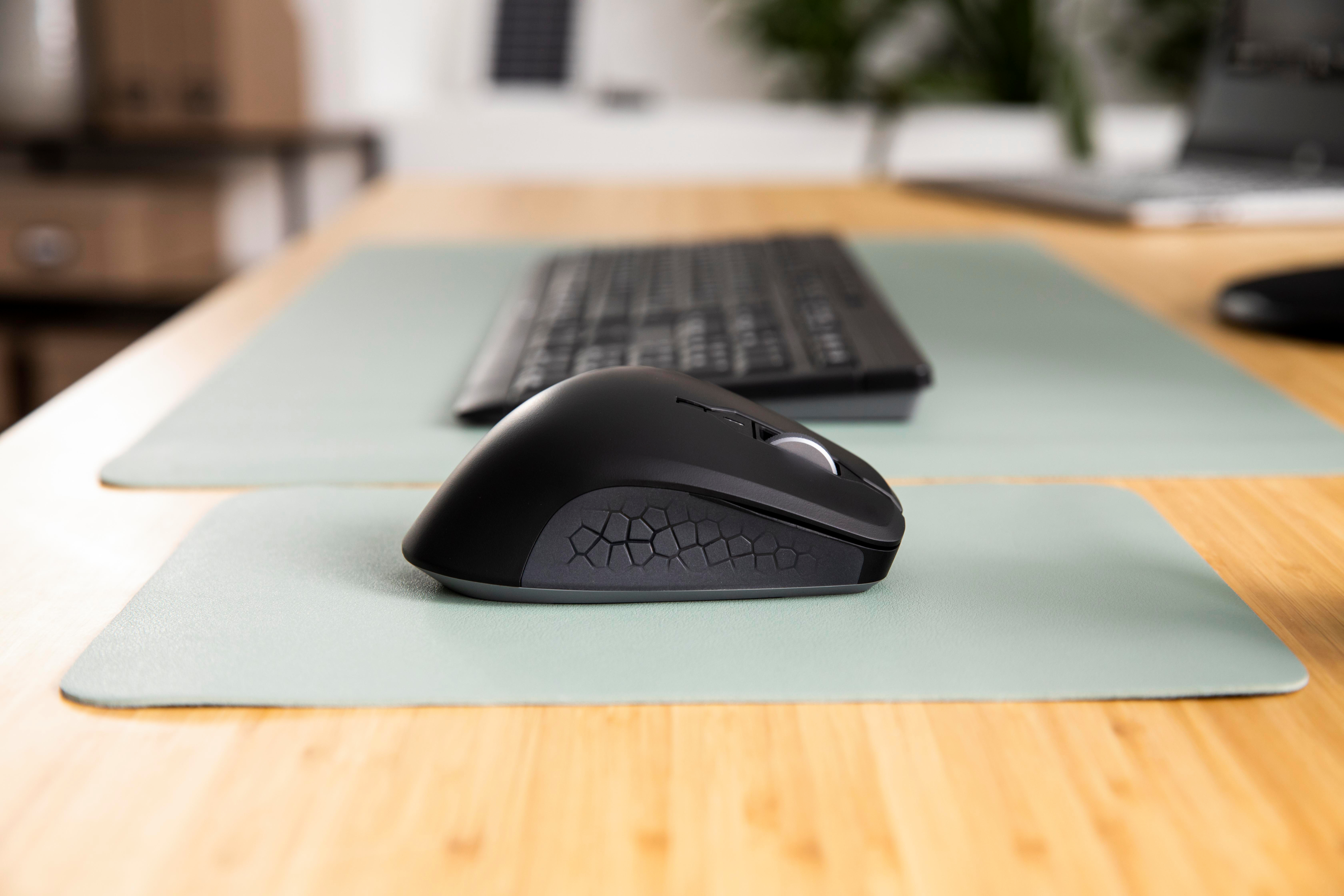 Schwarze Maus auf grüner Matte. Tastatur im Hintergrund. Holzschreibtisch. Die Szene ist gut beleuchtet.