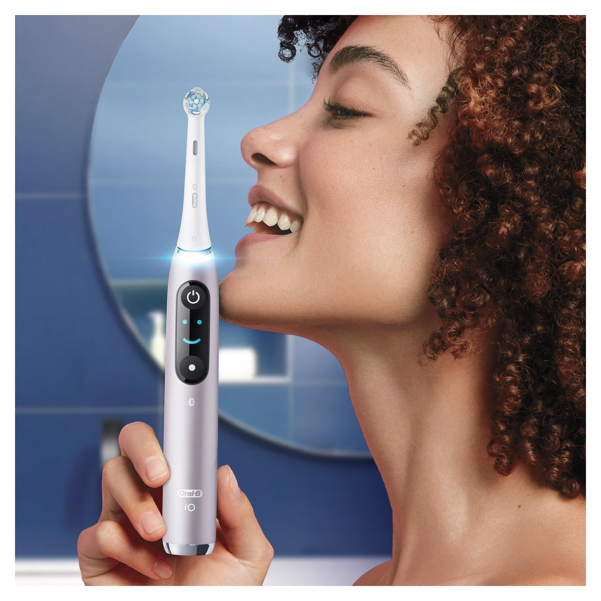 Kıvırcık saçlı, gülümseyen kadın, Oral-B iO diş fırçası kullanıyor. Arka plan mavi fayans duvar ve bir aynadır.