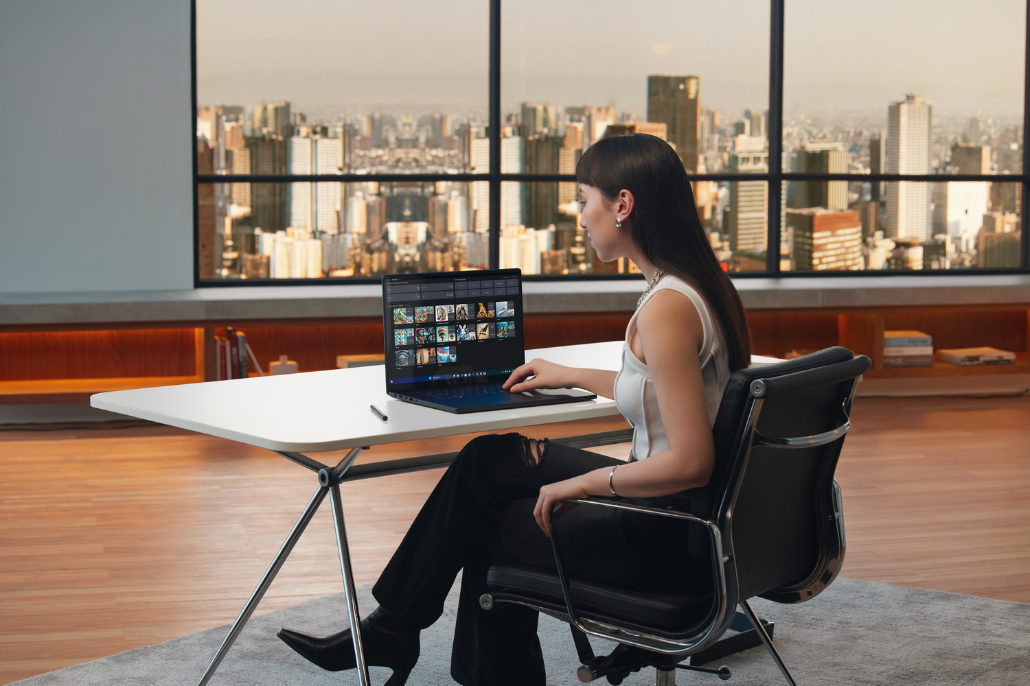 Mujer en escritorio con laptop. Vista de la ciudad de fondo. Lleva top blanco y pantalón negro.