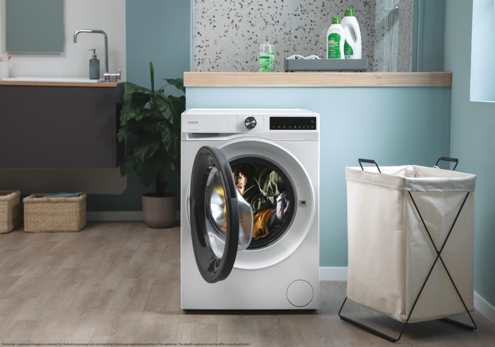 Une machine à laver blanche avec une porte ouverte, un panier à linge et des produits de nettoyage dans une salle de bain claire.