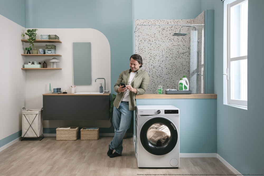 Homme dans salle de bain, adossé à machine à laver, regarde son téléphone. Pièce avec douche, lavabo et fenêtre.