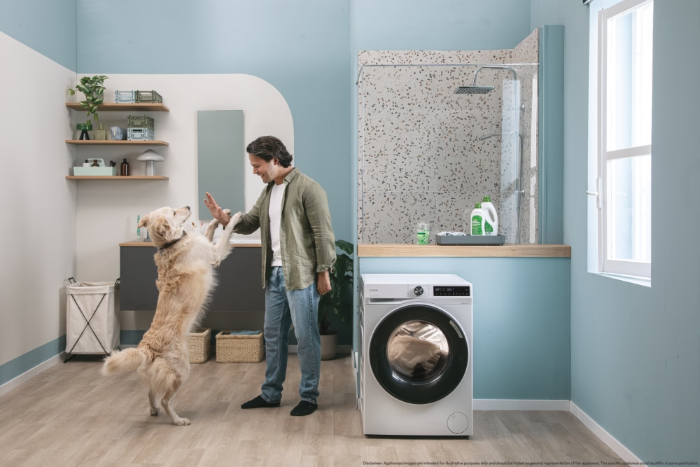 Homme dans une pièce avec un chien. Le chien se tient sur ses pattes arrières, tendant une patte. Lave-linge et douche en fond.