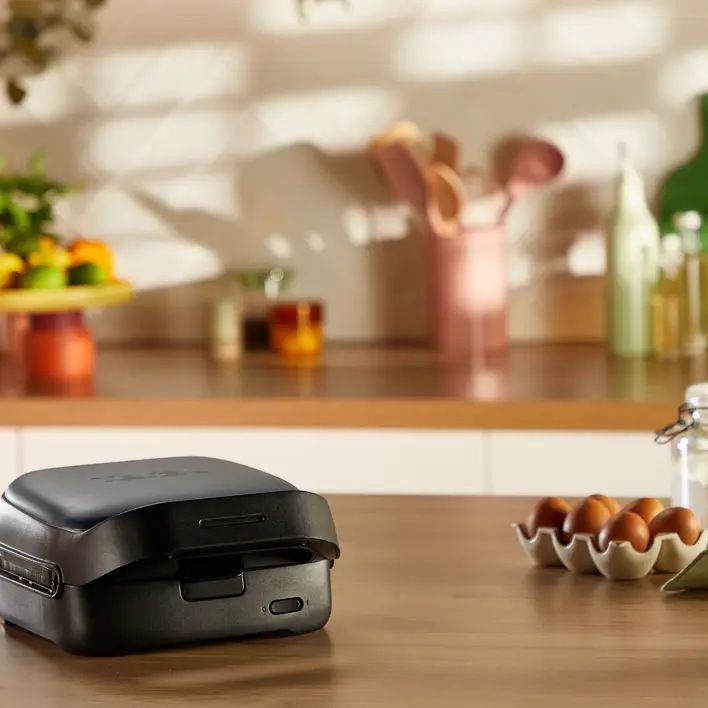 Czarny opiekacz do kanapek na drewnianym stole. Jajka, przybory i owoce w tle. Otoczenie kuchni.
