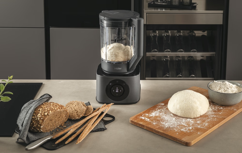 Scena di panificazione con macchina Haier, pane, pasta, farina e tagliere. I toni grigi e legno dominano la cucina.