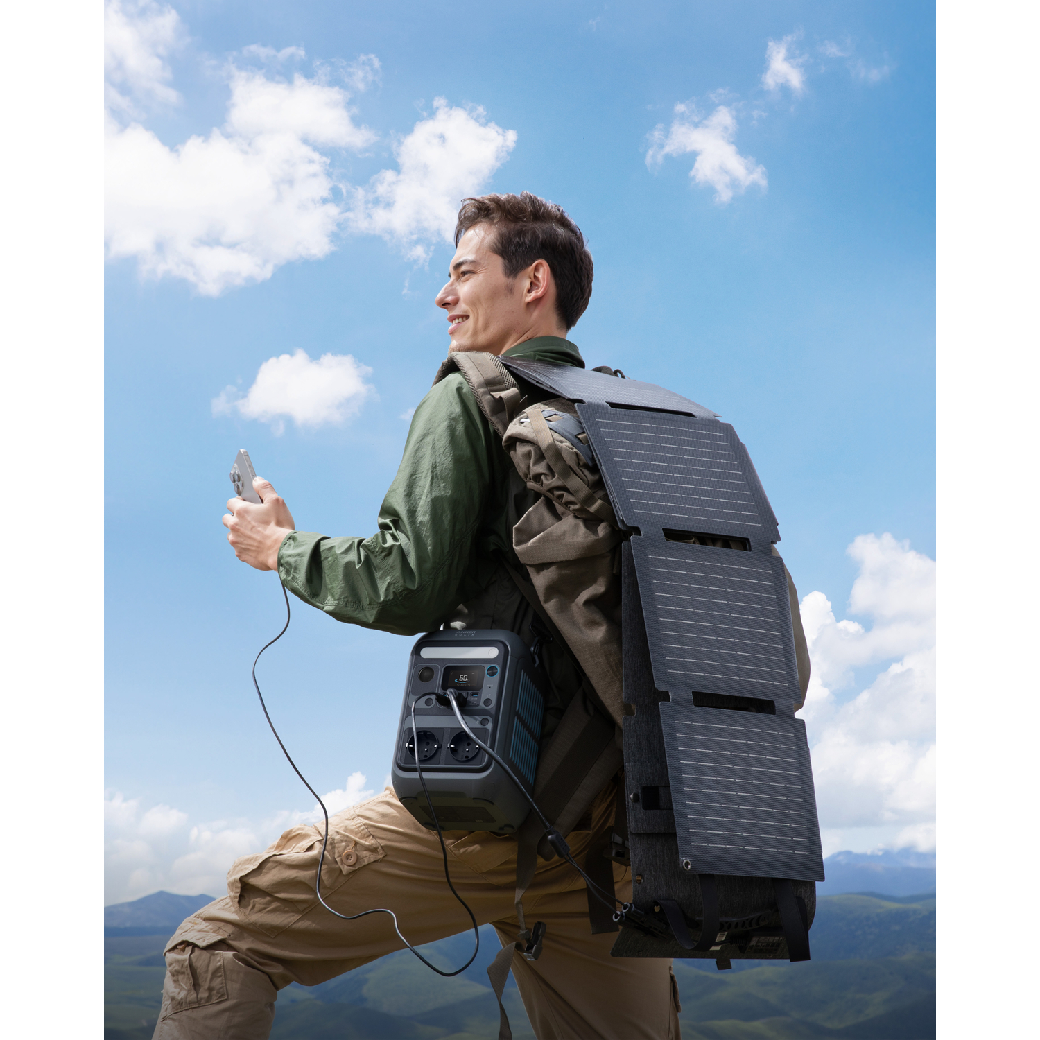 Homme avec sac à dos solaire, tenant téléphone, souriant, ciel bleu. Centrale électrique attachée. Vue montagnes.