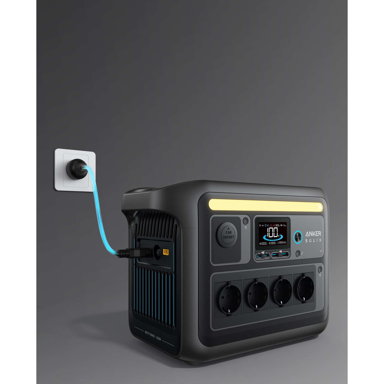 Black Anker Solix power station, plugged in, shows 100% charge.  White wall socket with blue cable. Grey background.