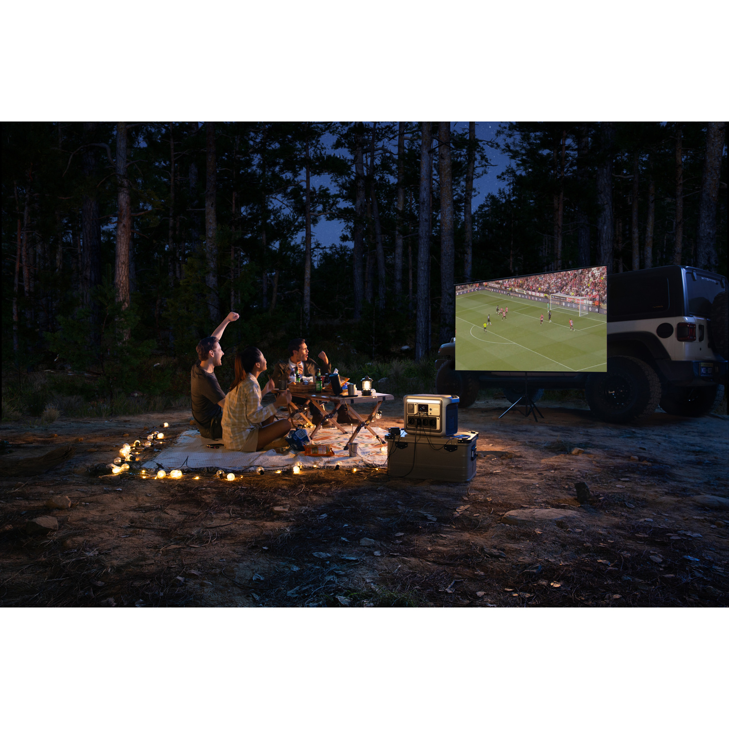 Group watches soccer on a screen in a forest at night. A jeep is parked nearby. String lights and a table are set up.