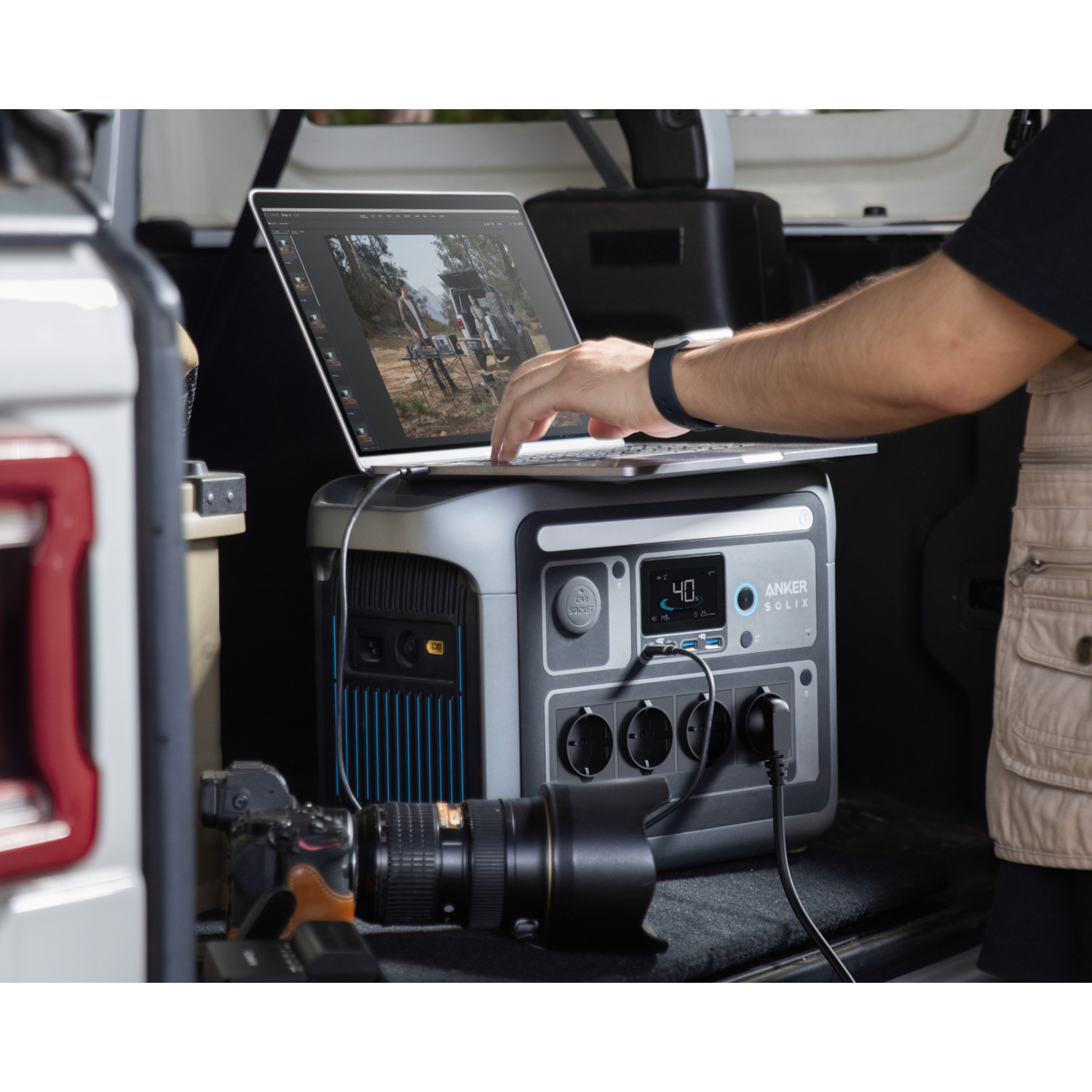 A person works on a laptop connected to a power station in a car. Camera and lens are in the foreground.