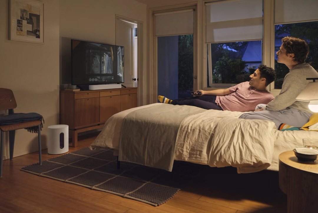 Dwóch mężczyzn na łóżku ogląda TV. Jeden trzyma pilota. Pokój ma TV, głośnik i okno z widokiem na zewnątrz.