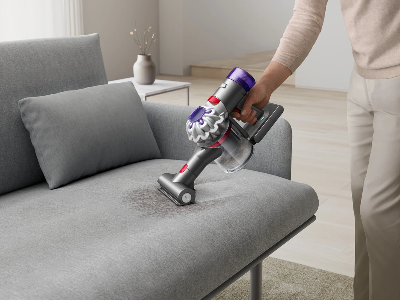 Egy személy egy szürke kanapét porszívózik egy vezeték nélküli Dyson porszívóval. A kanapén szürke párna és szőnyeg van, semleges háttér.