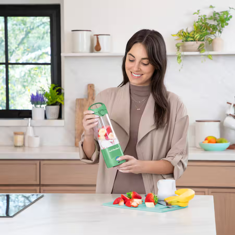 Kobieta w kuchni z zielonym blenderem. Truskawki, banany na blacie. Rośliny w tle.