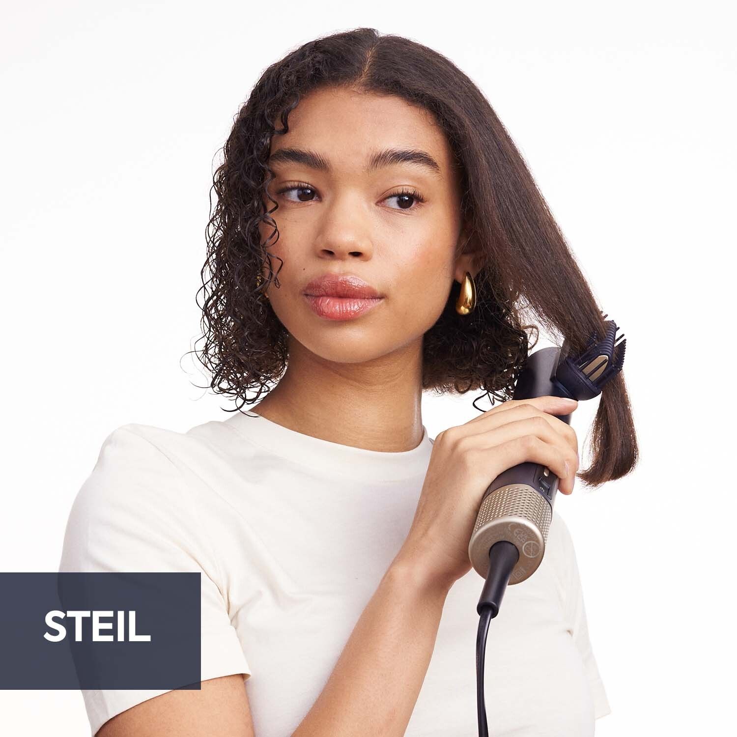 Vrouw gebruikt een stijltang. Ze draagt een wit shirt en gouden oorbellen. De achtergrond is wit.