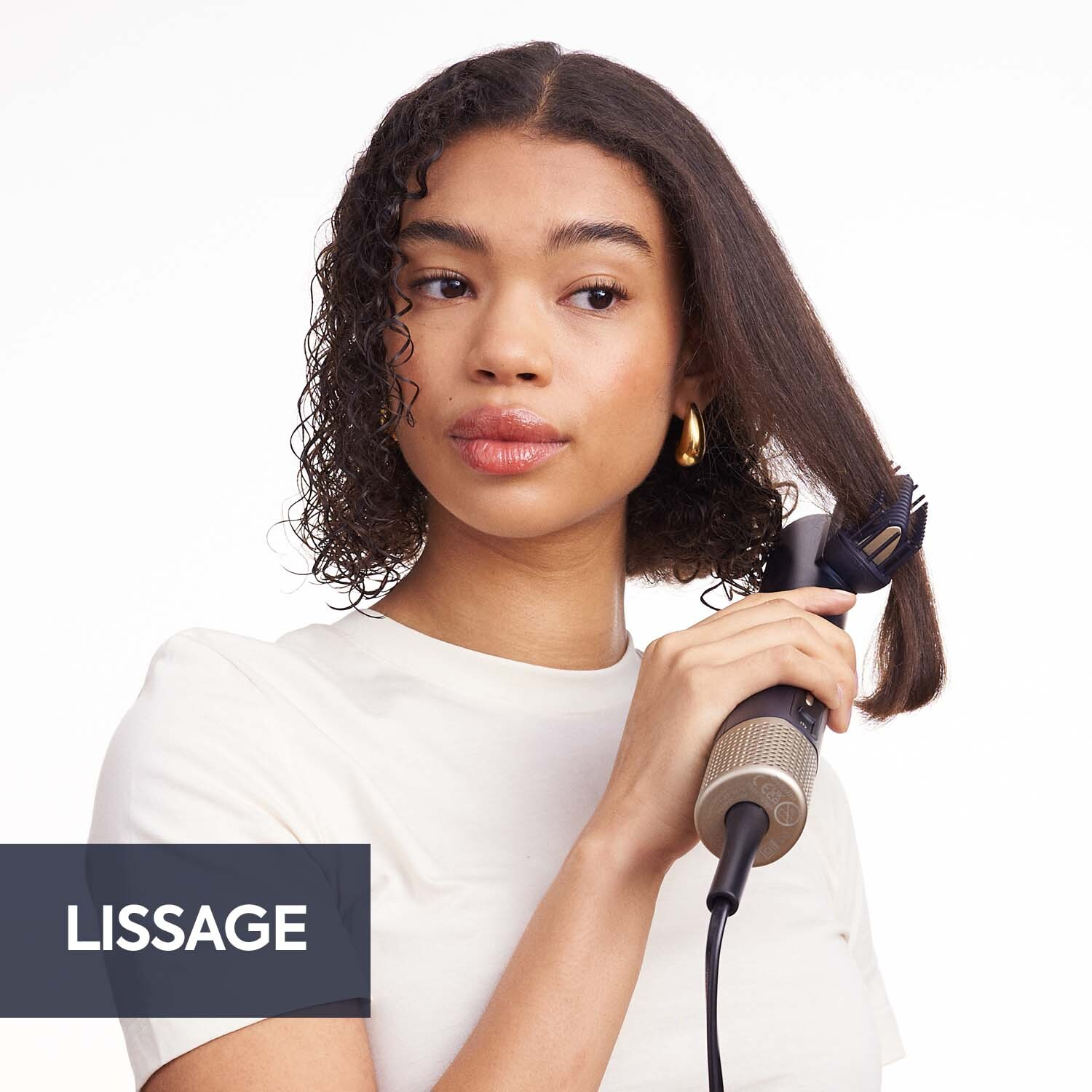 Une femme se lisse les cheveux avec un outil de coiffage. Elle porte un t-shirt blanc et des créoles dorées. Le fond est blanc.