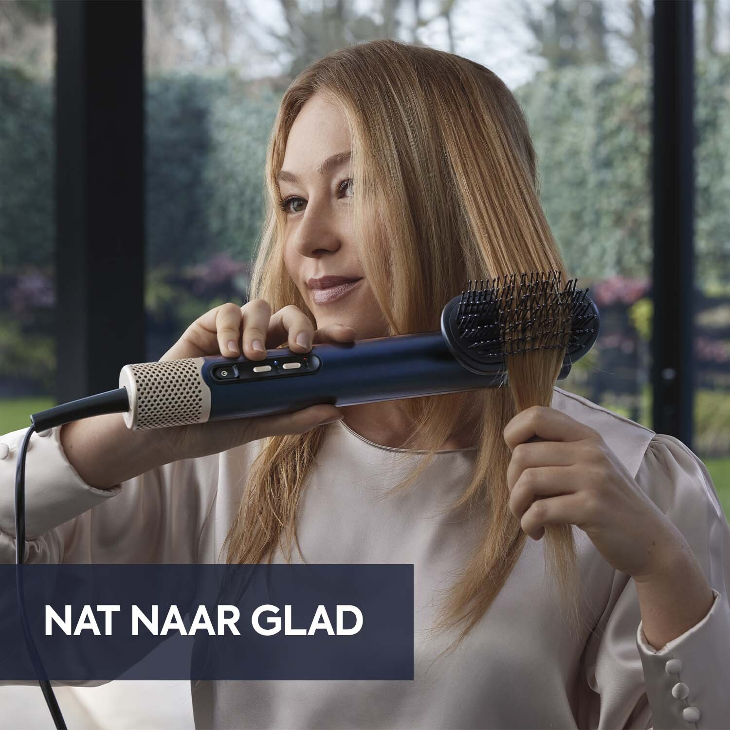 Vrouw die haar haar stylet met een blauw-witte föhn. Blond haar, wit overhemd. Achtergrond: groene bomen en ramen.