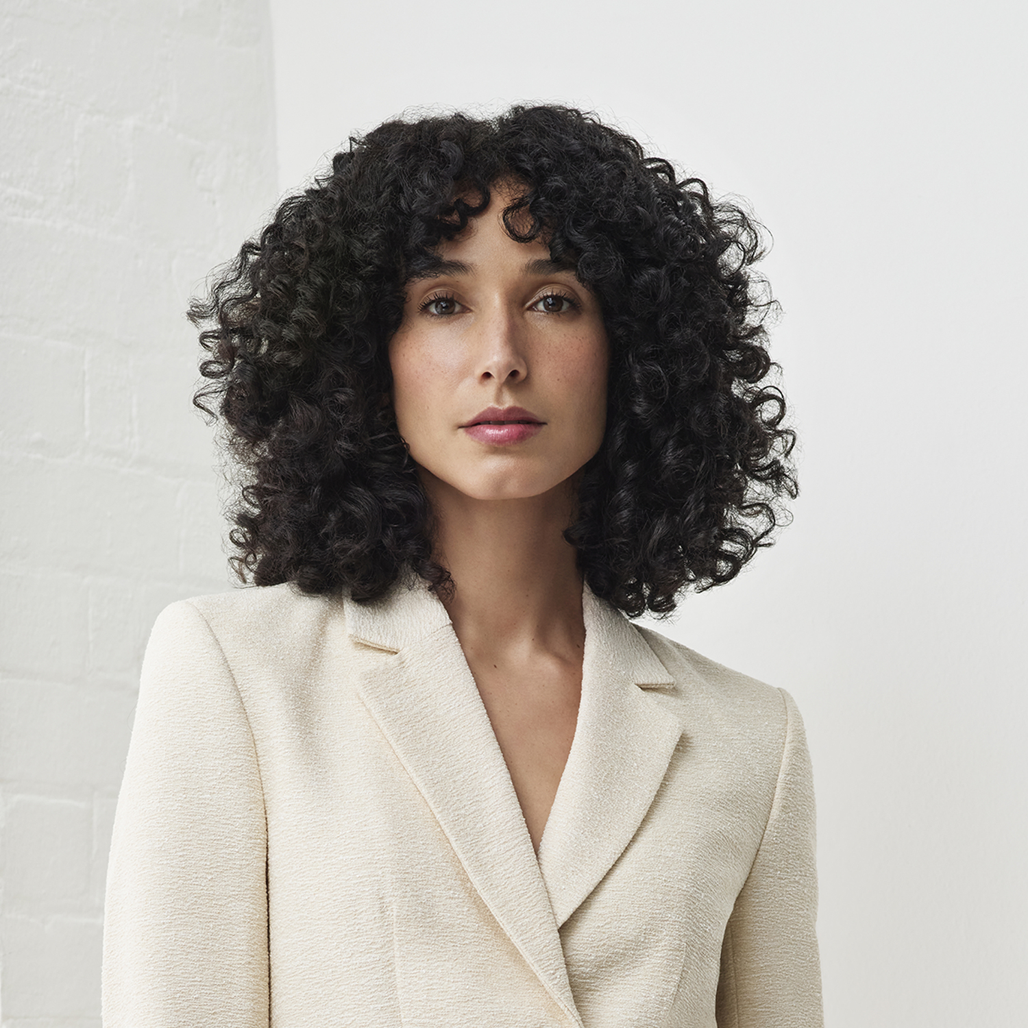 Woman with curly black hair and a cream blazer. White brick wall in background. She looks directly at the viewer.