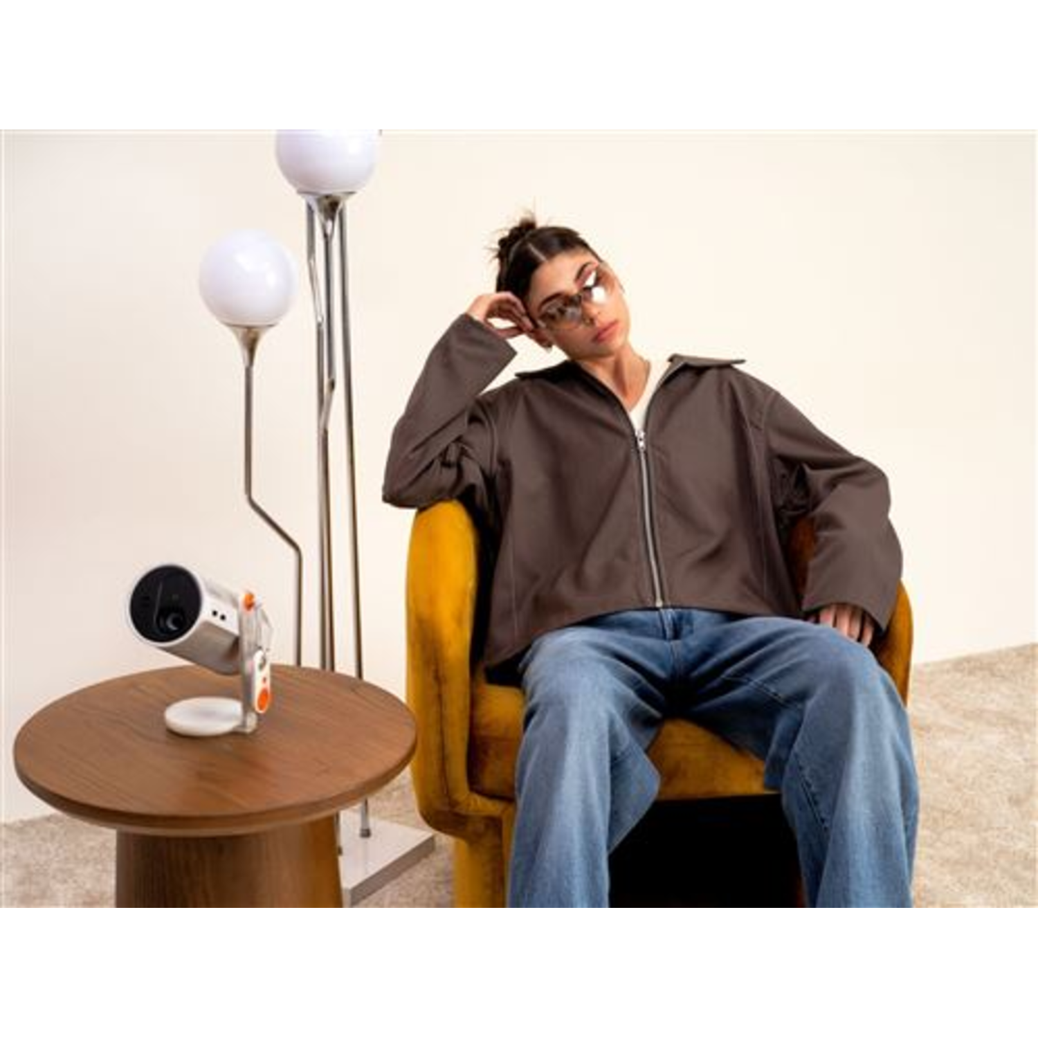 Woman in brown jacket and jeans, sunglasses, sits in a yellow chair. Lamps and a table with a camera are in the room.