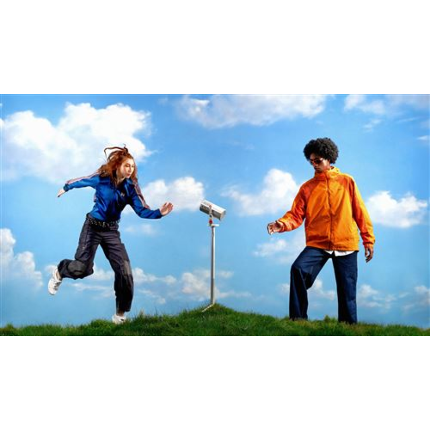 Woman in blue jacket running towards man in orange jacket. Both on grass, blue sky with clouds in background.