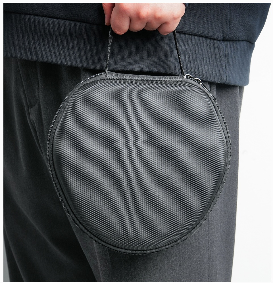 Osoba trzyma szarą, okrągłą, zapinaną na zamek walizkę z uchwytem. Walizka wydaje się być na słuchawki. Osoba ma na sobie ciemne ubrania.