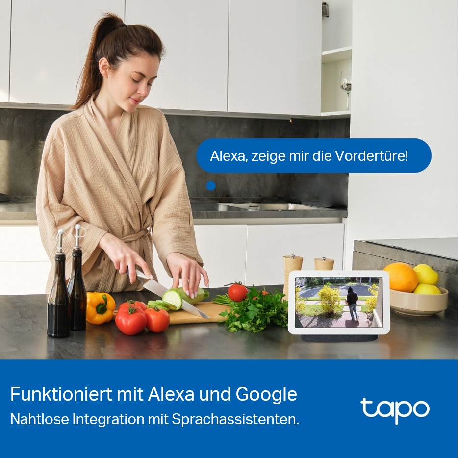 Frau schneidet Gemüse in der Küche. Smart Display zeigt die Haustür. Gemüse und Flaschen auf der Theke. Text: Alexa, Google.