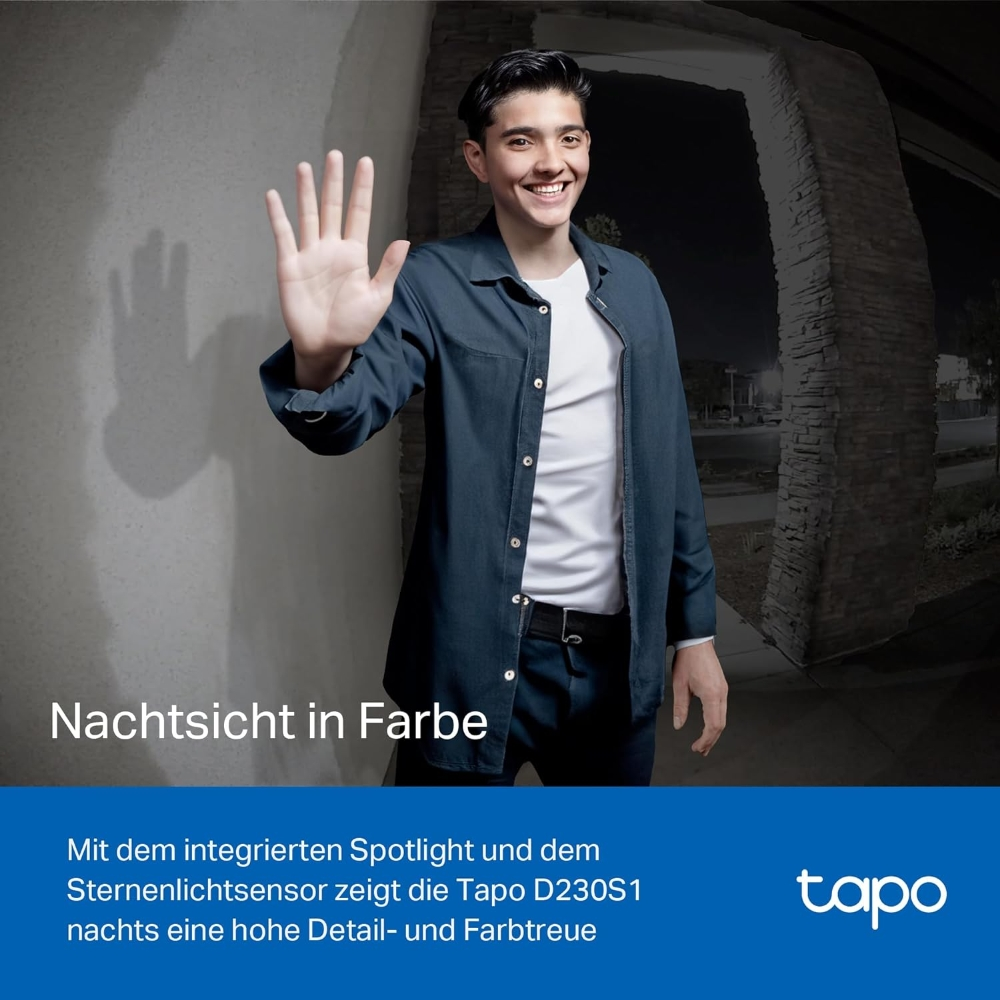 Ein lächelnder Mann winkt mit der Hand und steht nachts vor einem Gebäude. Er trägt ein blaues Hemd über einem weißen T-Shirt.