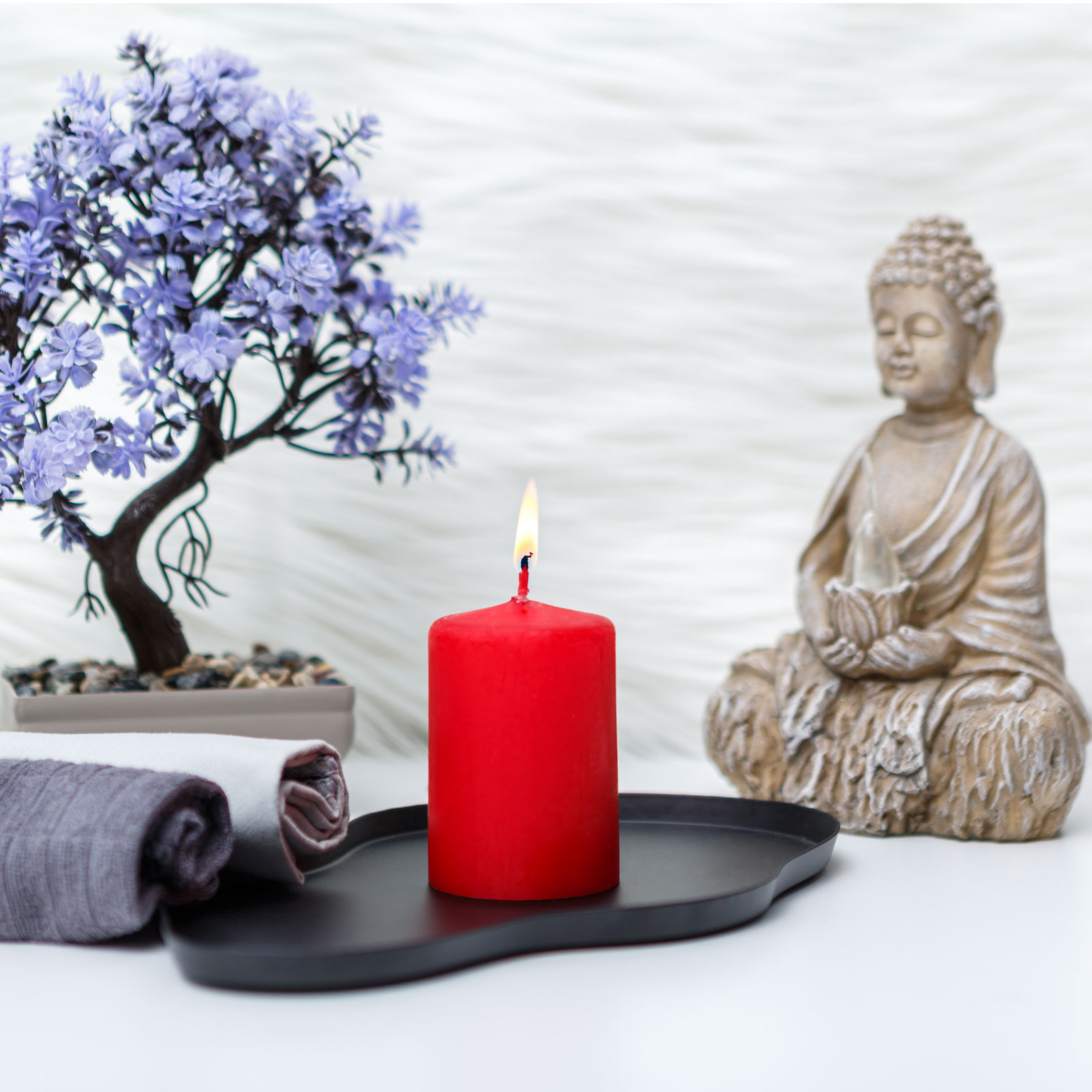 Piros gyertya fekete tálcán, Buddha szobor, bonsai fa és hajtogatott törölközők. Fehér, bolyhos háttér.