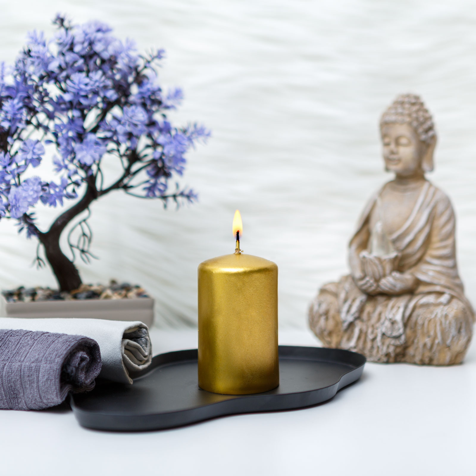 Egy arany gyertya lánggal egy fekete tálcán áll. A háttérben Buddha-szobor és lila fa látható.