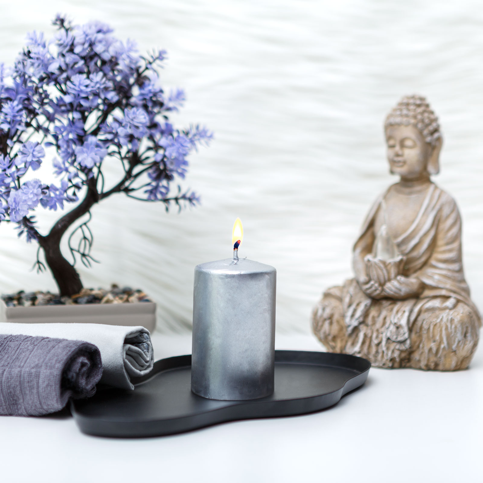 Egy meggyújtott ezüst gyertya fekete tálcán. Buddha szobor és lila fa a háttérben. Összetekert törölközők az előtérben.