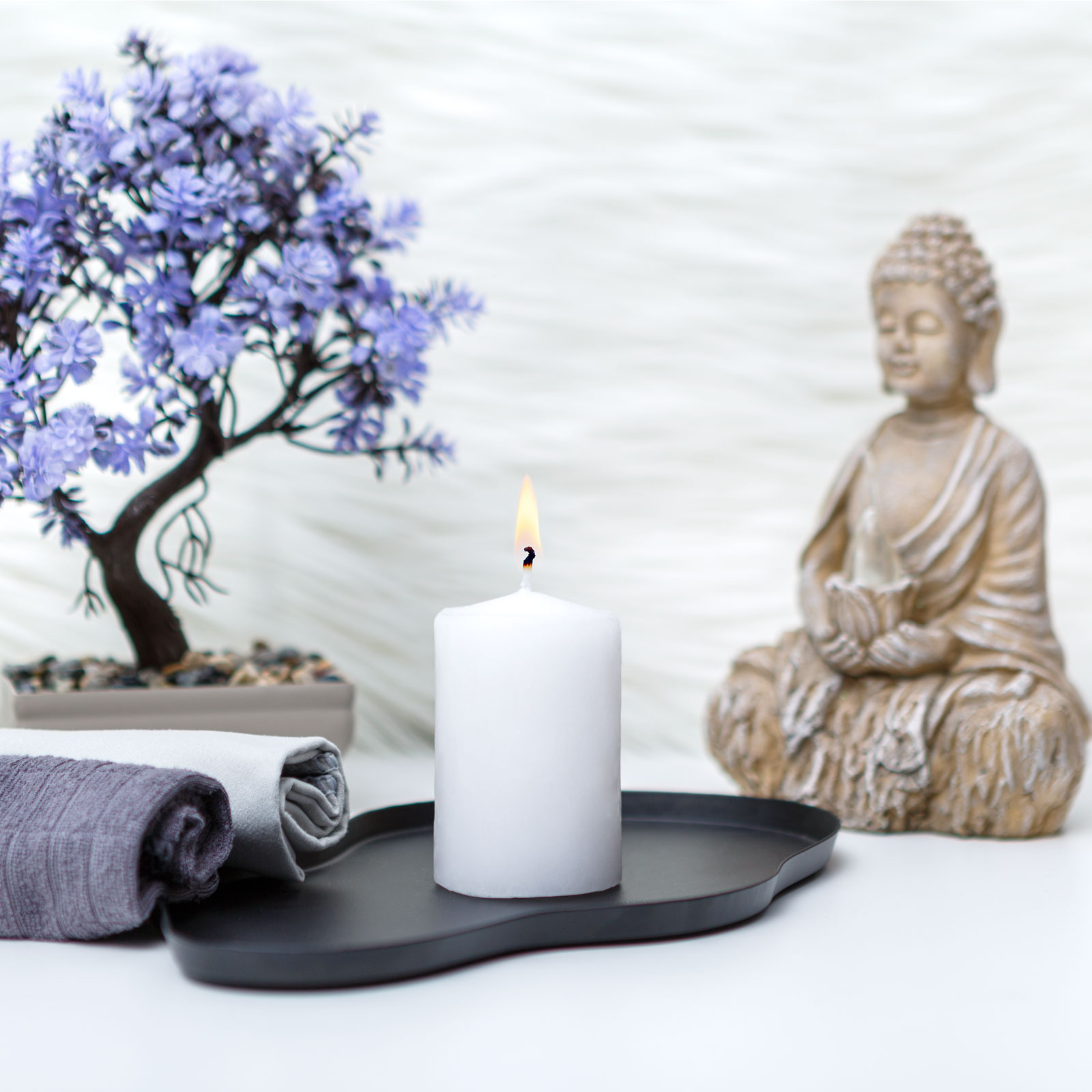 Egy meggyújtott fehér gyertya fekete tálcán. Buddha szobor, lila fa és feltekert törölközők a háttérben.