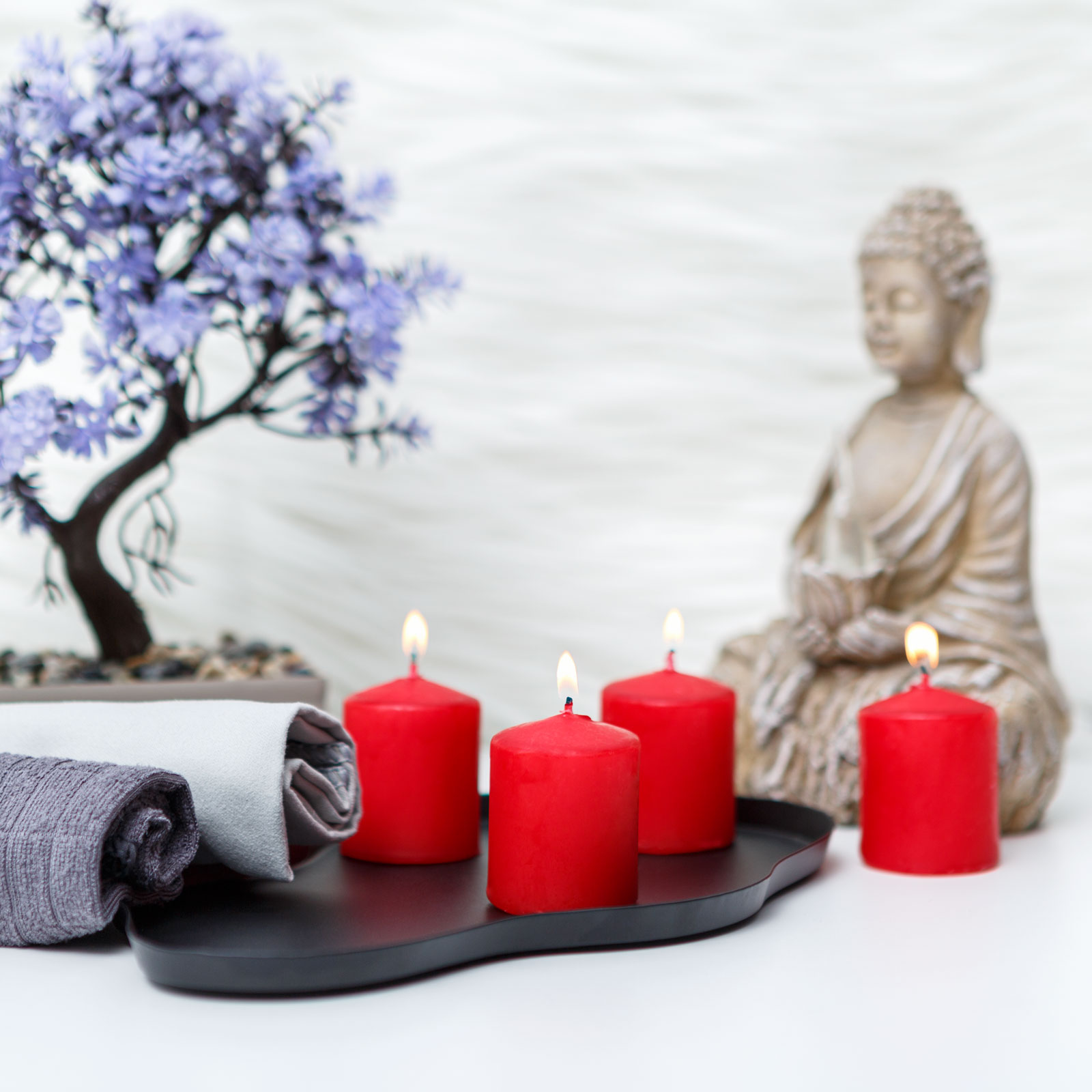 Piros gyertyák fekete tálcán, törölközők, bonsai fa és Buddha szobor fehér felületen.